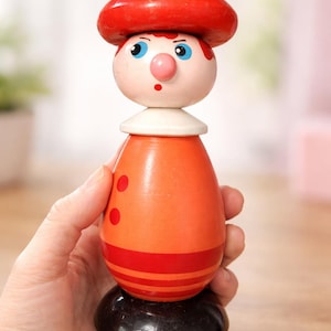 Puede incluir: Figura de juguete de madera con sombrero rojo y cuerpo naranja. La figura tiene cuello blanco, ojos azules y nariz rosa. El cuerpo es naranja con rayas y puntos rojos. La base es negra.