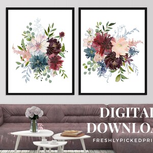 May include: Two framed floral art prints featuring watercolor bouquets in shades of burgundy, teal, and blush pink. The bouquets have gold accents and are set against a white background. The prints are displayed above a mauve sofa with white side tables.