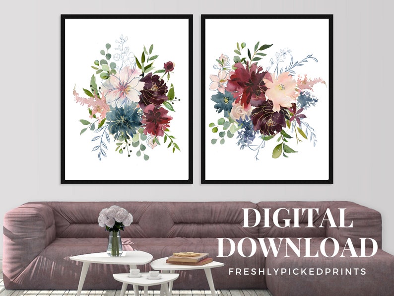 May include: Two framed floral art prints featuring watercolor bouquets in shades of burgundy, teal, and blush pink. The bouquets have gold accents and are set against a white background. The prints are displayed above a mauve sofa with white side tables.