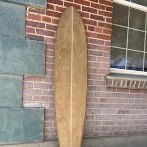 May include: A wooden surfboard with a light brown finish and a thin, lighter-colored stripe down the center. The surfboard is leaning against a brick wall. The board's shape is long and narrow, designed for riding waves.