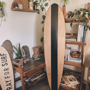 May include: A wooden surfboard with a black stripe down the center. The surfboard is leaning against a wooden shelf with books and plants. The text "Pray for Surf" is on a wooden sign in the foreground.