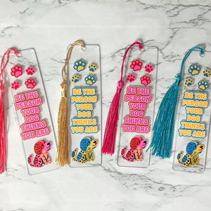 May include: Four clear rectangular acrylic bookmarks, each featuring a colorful illustration of a dog, paw prints, and the text "BE THE PERSON YOUR DOG THINKS YOU ARE." Each bookmark is finished with a decorative silk tassel in pink, gold, or blue.