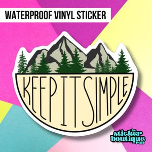 May include: A white and yellow vinyl sticker with a black outline. The sticker features a mountain range with green trees in front of the mountains. The text "KEEP IT SIMPLE" is written in black on the sticker.