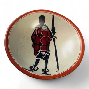 May include: A small, round, white stone bowl with a red rim. The bowl features a black and red silhouette of a person holding a spear.