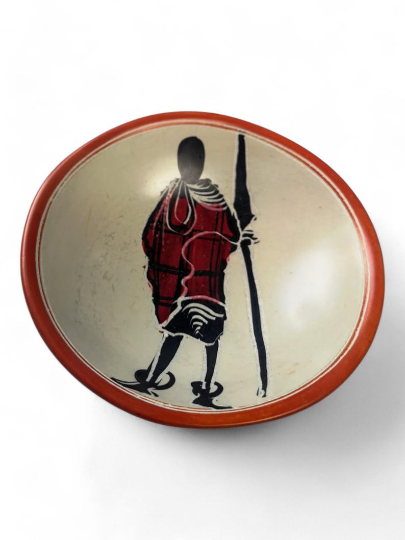 May include: A small, round, white stone bowl with a red rim. The bowl features a black and red silhouette of a person holding a spear.