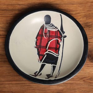 May include: A white ceramic plate with a black rim features a hand-painted design of a person in a red and black plaid shirt holding a spear. The plate is a decorative piece with a traditional African design.