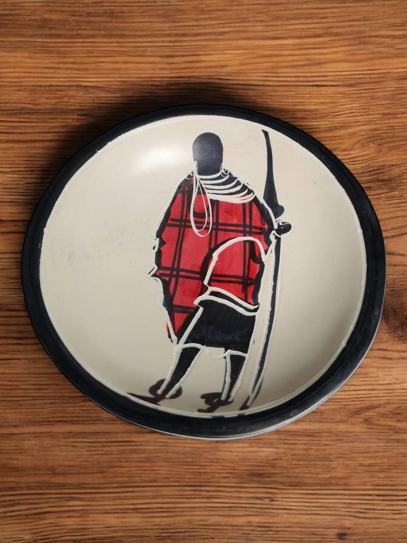 May include: A white ceramic plate with a black rim features a hand-painted design of a person in a red and black plaid shirt holding a spear. The plate is a decorative piece with a traditional African design.