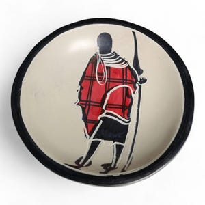 May include: A round, white ceramic bowl with a black rim. The bowl features a hand-painted design of a person in a red and black patterned robe, holding a spear.