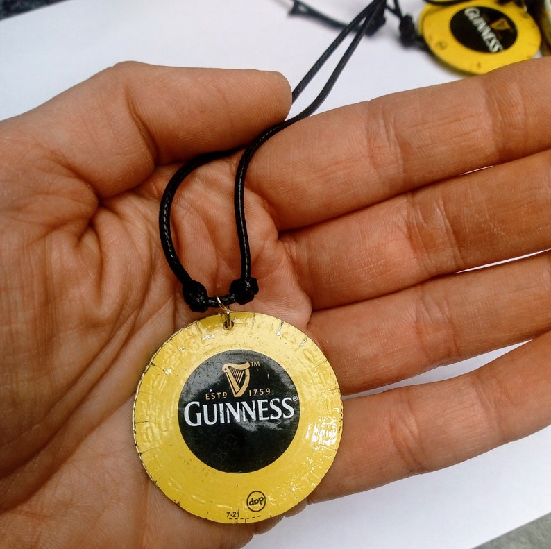 May include: A black cord necklace with a round, yellow and black Guinness beer can lid pendant. The pendant features the Guinness logo and the text "ESTD 1759 GUINNESS" in black.
