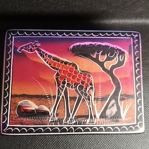 May include: A black wooden box with a decorative border and a colourful image of a giraffe standing in a savanna landscape with a tree in the background. The giraffe is red, orange, and white, and the background is a sunset gradient of orange, pink, and purple.