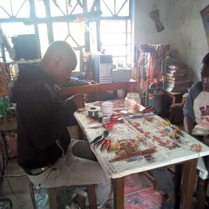 May include: A person is working on a table with a variety of beads, wire, and tools. The table is covered in a white cloth with a newspaper underneath. There are several strands of beads hanging on a rack in the background.
