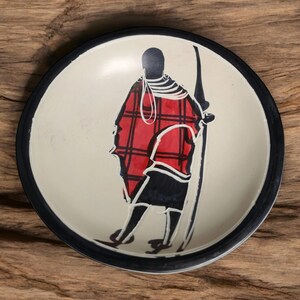 May include: A white ceramic dish with a black rim features a hand-painted design of a person in a red and black plaid robe holding a spear. The background is white.