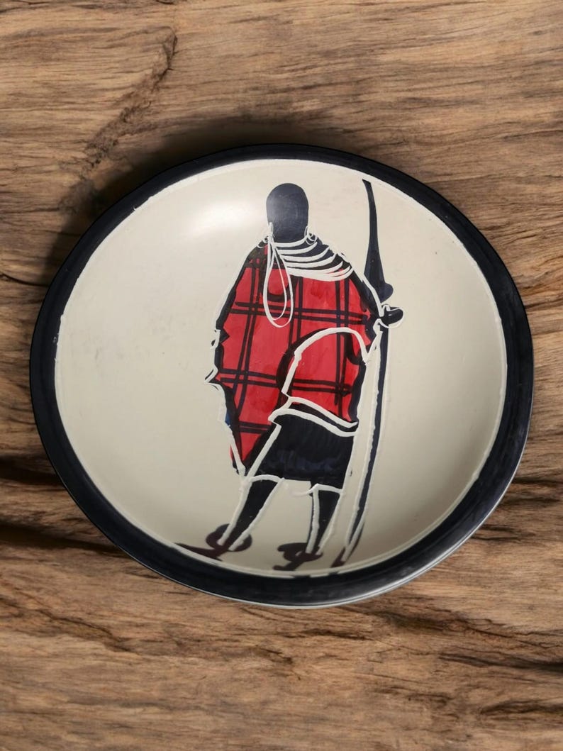 May include: A white ceramic dish with a black rim features a hand-painted design of a person in a red and black plaid robe holding a spear. The background is white.
