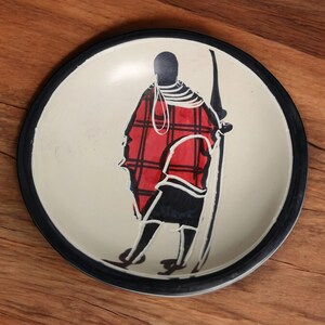 May include: A white ceramic plate with a black rim features a hand-painted silhouette of a person wearing a red and black plaid garment. The person is holding a spear and is standing against a white background.