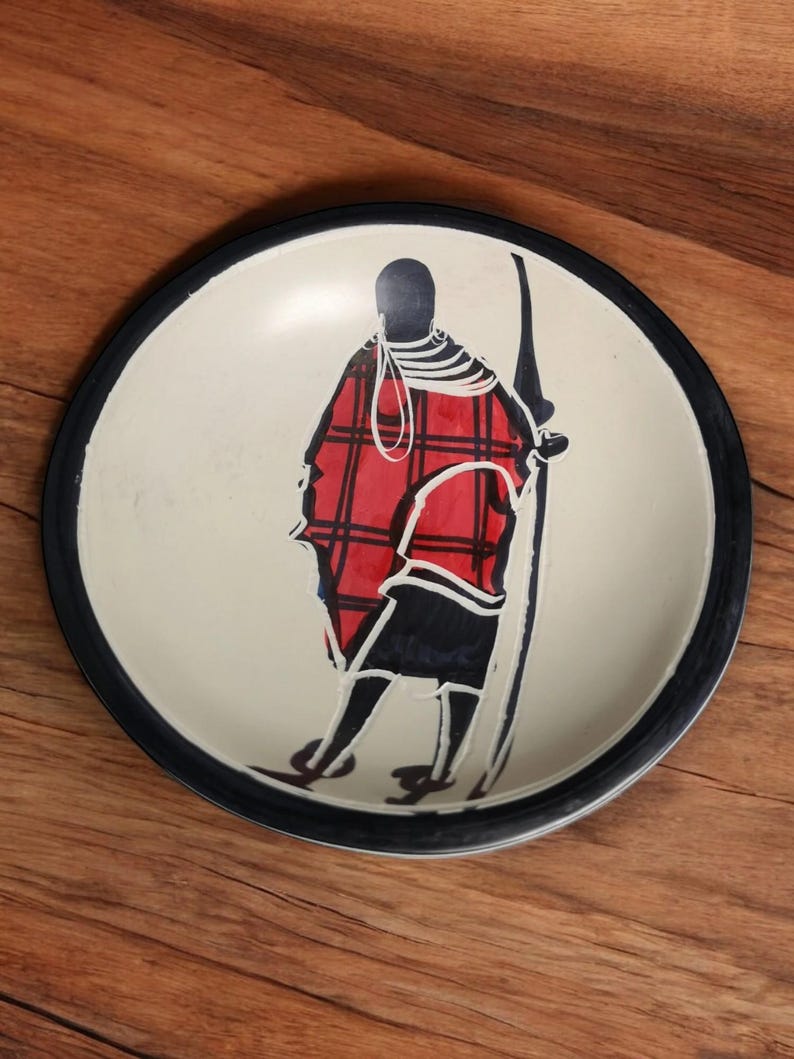 May include: A white ceramic plate with a black rim features a hand-painted silhouette of a person wearing a red and black plaid garment. The person is holding a spear and is standing against a white background.