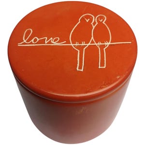 Puede incluir: Contenedor redondo de esteatita naranja con tapa. La tapa tiene un diseño blanco de dos pájaros posados en un alambre con la palabra "love" escrita en el alambre.