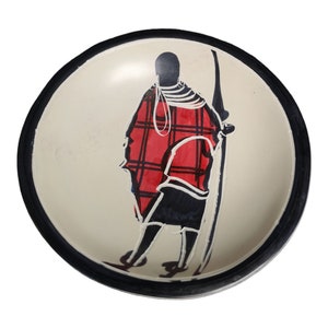 May include: A white ceramic bowl with a black rim and a hand-painted design of a person in a red and black patterned robe. The person is holding a spear and is standing in a profile view.
