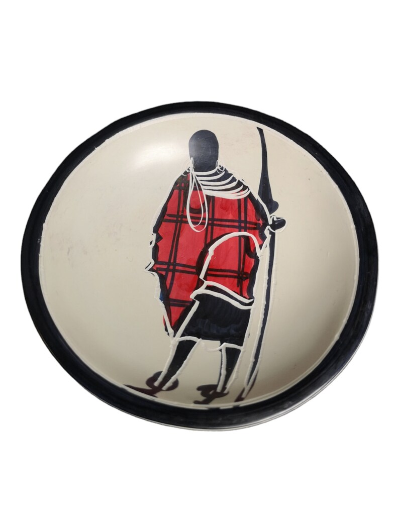 May include: A white ceramic bowl with a black rim and a hand-painted design of a person in a red and black patterned robe. The person is holding a spear and is standing in a profile view.