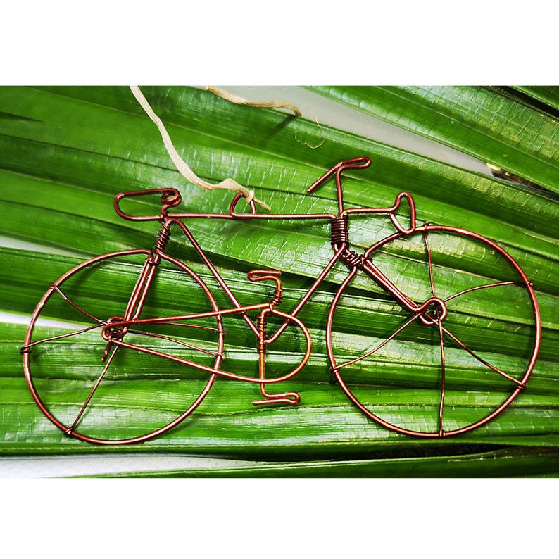 May include: A copper wire bicycle sculpture with a brown twine string attached to the handlebars. The sculpture is resting on a green leaf.