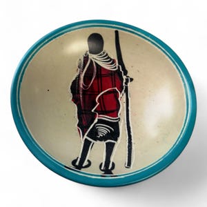 May include: A small, round, teal-colored soapstone bowl with a hand-painted design of a person in a red and black patterned robe. The bowl is perfect for holding jewelry, keys, or other small items.