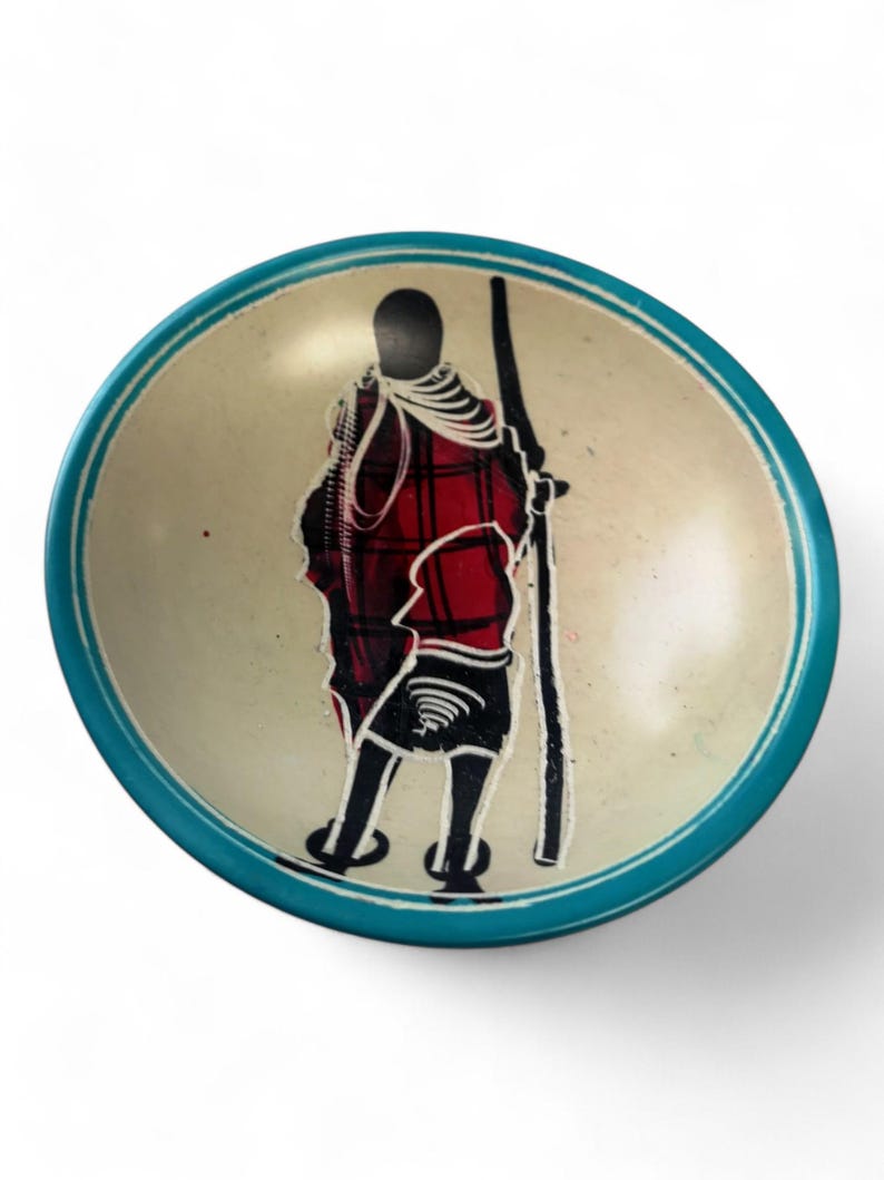 May include: A small, round, teal-colored soapstone bowl with a hand-painted design of a person in a red and black patterned robe. The bowl is perfect for holding jewelry, keys, or other small items.