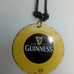 May include: A round, yellow metal keychain with a black and white Guinness logo. The logo features a harp, the words "ESTD 1759", and the word "GUINNESS" in a black font. The keychain has a black cord attached to it.