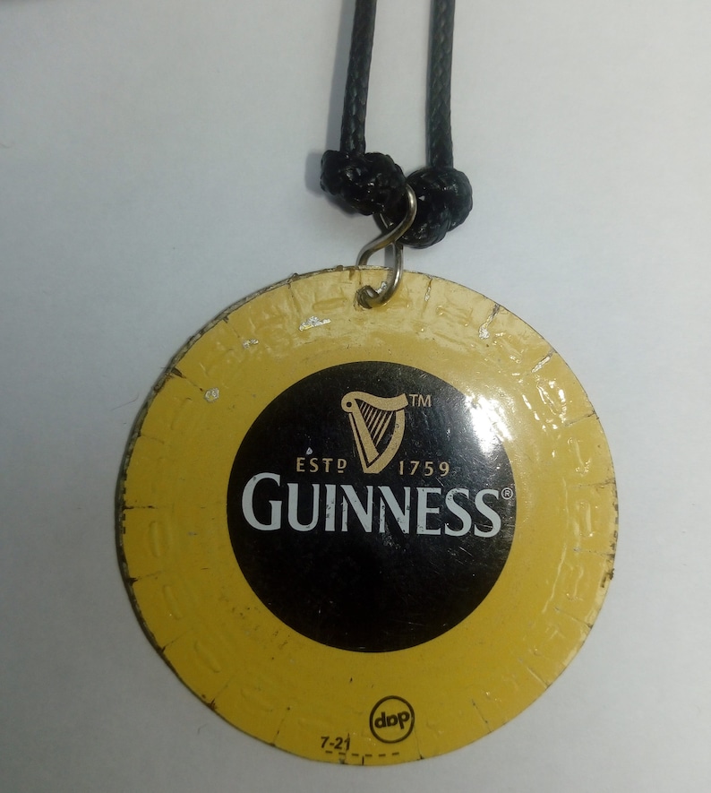May include: A round, yellow metal keychain with a black and white Guinness logo. The logo features a harp, the words "ESTD 1759", and the word "GUINNESS" in a black font. The keychain has a black cord attached to it.
