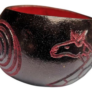 May include: A small, round, black bowl with a red interior. The bowl has a red spiral design on one side and a red stylised animal design on the other side.