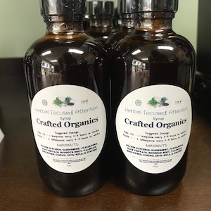 Peut inclure: Deux bouteilles en verre brun de sirop d'attention concentrée aux herbes de Crafted Organics. Les étiquettes sont blanches avec des accents verts et noirs. Les bouteilles sont sur une surface en bois.