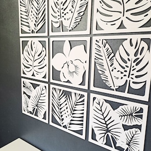 May include: A set of nine white wooden wall decor panels featuring various tropical leaf designs. The panels are arranged in a grid pattern and are all the same size.
