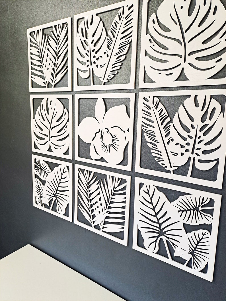 May include: A set of nine white wooden wall decor panels featuring various tropical leaf designs. The panels are arranged in a grid pattern and are all the same size.