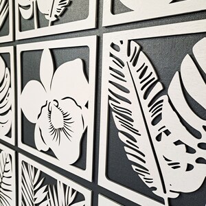 May include: A set of nine white wooden wall decor panels featuring various tropical leaf and flower designs. The panels are arranged in a grid pattern and are set against a dark gray background.