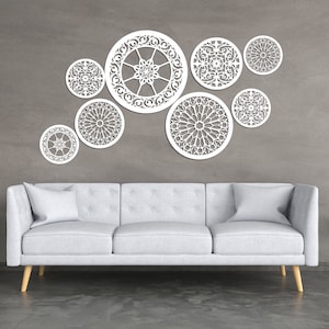 Könnte beinhalten: Ein weißes Sofa mit zwei Kissen steht vor einer grauen Wand mit weißer dekorativer Wandkunst. Die Wandkunst besteht aus acht runden Teilen mit komplizierten Designs. Es gibt zwei kleine schwarze Beistelltische mit einer Pflanze und einer Tasse auf jedem.