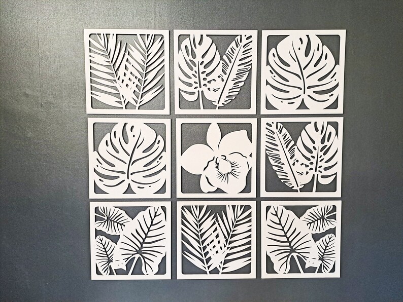 May include: Nine white square wall decor pieces with cut-out tropical leaf designs. The designs include palm leaves, monstera leaves, and an orchid flower.