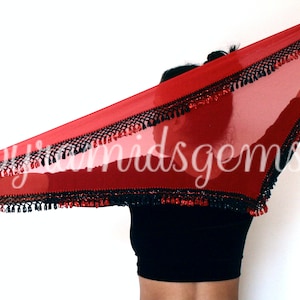 Puede incluir: Un pañuelo de danza rojo transparente con flecos negros y cuentas rojas. El pañuelo está sostenido por la mano de una persona.