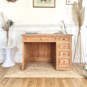 Op de afbeelding: Een lichtbruine houten bureau met een enkele lade en meerdere laden aan de rechterkant. Het bureau staat op een jute vloerkleed, met een ingelijste bloemenprint en een gedroogde krans aan de muur. Een discobal en decoratief pampasgras zijn ook zichtbaar.
