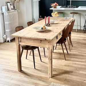 Puede incluir: Una mesa de comedor rectangular de madera clara, con un acabado envejecido, rodeada de sillas de madera. La mesa está decorada con un plato decorativo y velas. La habitación tiene suelo de parquet y un radiador.