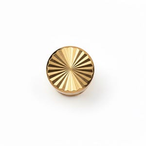 Puede incluir: Objeto circular dorado con un diseño de rayos de sol en la parte superior. El objeto tiene un borde estriado y una base plana. La parte superior presenta líneas radiales, creando un patrón texturizado. El objeto está sobre un fondo blanco. Diámetro aproximado de 3 cm.