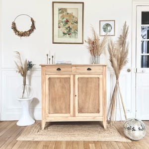 Op de afbeelding: Een lichtgekleurde houten kast met twee lades en twee deuren. De kast staat tegen een witte muur, met een ingelijste bloemenprint en een krans van gedroogde bloemen. Een jute vloerkleed en een discobal liggen op de houten vloer.
