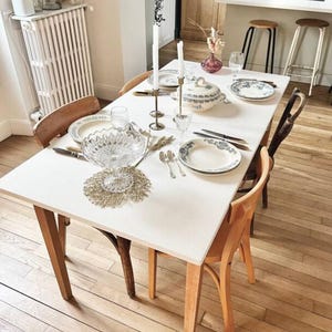 Könnte beinhalten: Ein weißer Tisch mit einer weißen Tischdecke, der für ein Abendessen gedeckt ist. Der Tisch ist mit zwei Gedecken gedeckt, jedes mit einem weißen Teller, einem Glas und Besteck. In der Mitte des Tisches steht eine Schüssel mit Obst und zwei Kerzenständer mit weißen Kerzen.