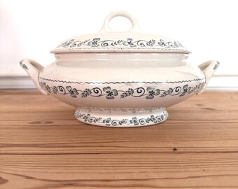 May include: Cream-colored ceramic serving dish with a lid and handles. The dish is decorated with a blue floral pattern around the rim, body, and base. The lid has a loop handle. The dish is on a wooden surface.