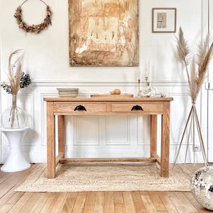 Op de afbeelding: Een lichte houten consoletafel met twee laden en zwarte metalen handgrepen. De tafel staat in een kamer met witte muren en een lichte houten vloer. Er ligt een tapijt onder de tafel en aan de muur erachter hangt een groot abstract schilderij.