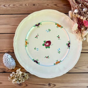 Peut inclure: Grande assiette en céramique blanc cassé à bord festonné. Décor floral peint à la main avec une rose bordeaux centrale et des fleurs orange, violettes et bleues. Un anneau bleu clair entoure le motif floral.