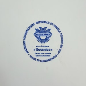 May include: A white porcelain plate with a blue circular emblem in the center. The emblem features text and a decorative design, including the words "VILLEROY & BOCH" and "Botanica". The plate is marked "MADE IN LUXEMBOURG".
