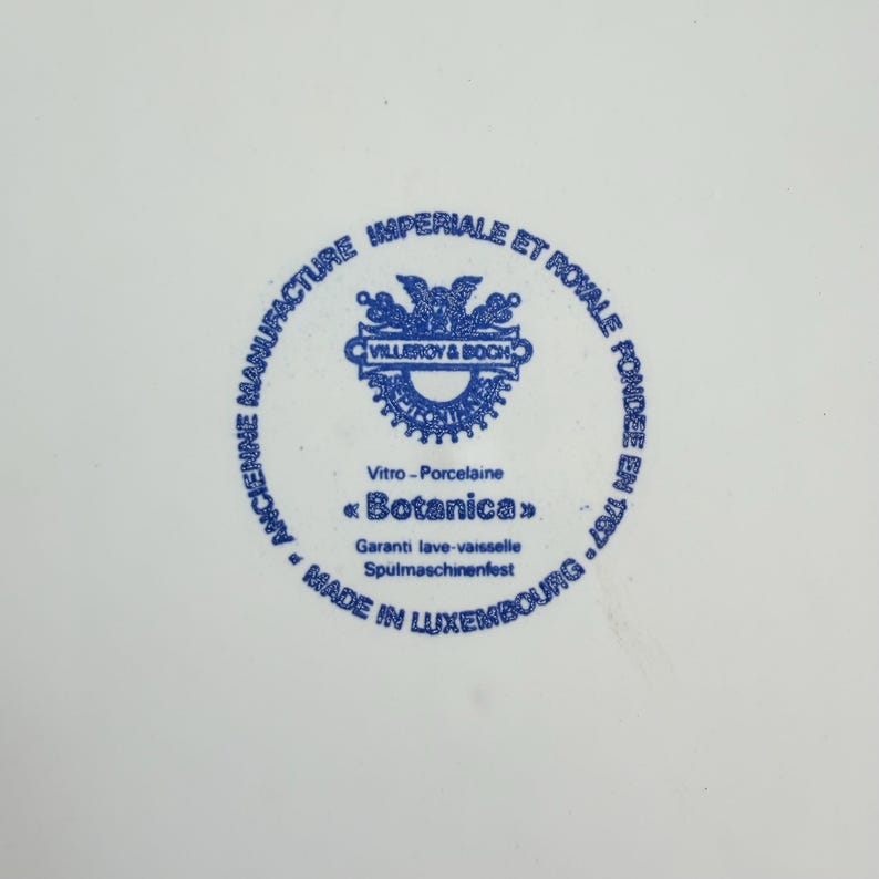 May include: A white porcelain plate with a blue circular emblem in the center. The emblem features text and a decorative design, including the words "VILLEROY & BOCH" and "Botanica". The plate is marked "MADE IN LUXEMBOURG".