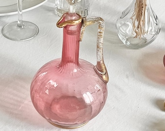 Jarra antigua de cristal soplado rosa con asa dorada y tapón facetado, elegancia vintage y encanto bohemio.