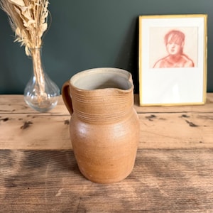May include: A tan ceramic pitcher with a smooth, rounded body and a textured neck featuring horizontal ridges. The pitcher has a single side handle and a wide, open spout. The interior of the vessel is a light, neutral off-white color.