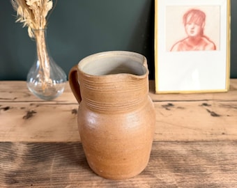 May include: A tan ceramic pitcher with a smooth, rounded body and a textured neck featuring horizontal ridges. The pitcher has a single side handle and a wide, open spout. The interior of the vessel is a light, neutral off-white color.
