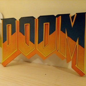 Peut inclure: Une enseigne en bois avec le mot "DOOM" en 3D. Les lettres sont peintes dans un dégradé d'orange et de jaune, avec un contour bleu foncé.