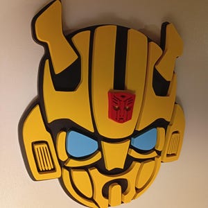 Peut inclure: Décoration murale en forme de tête de Bumblebee jaune et noir, avec des yeux bleus et un symbole Autobot rouge. L'œuvre d'art en 3D est montée sur un mur blanc et présente un design en couches.